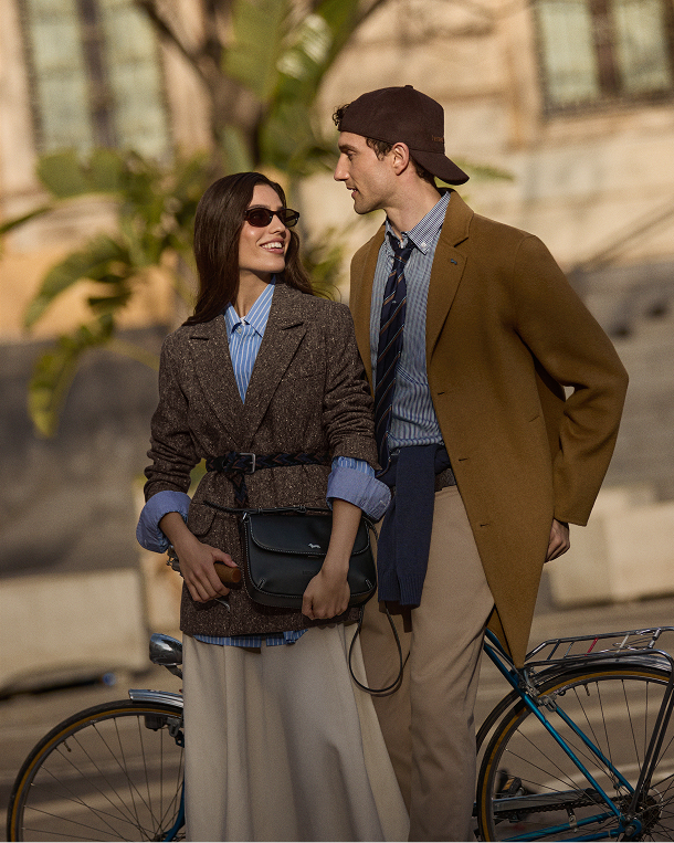 Campaign image for the Harmont & Blaine FW26 collection, themed Reinventing Daywear. A smiling couple looks at each other outdoors in a sunny urban setting, with a bicycle in the foreground. The woman wears a dark brown tweed double-breasted blazer cinched at the waist with a belt over a blue and white striped shirt, paired with a long beige pleated skirt. She wears dark sunglasses and carries a black leather crossbody bag. The man, wearing a brown baseball cap, sports a camel coat, a blue and white checkered shirt, and a dark striped tie, along with beige chino trousers.